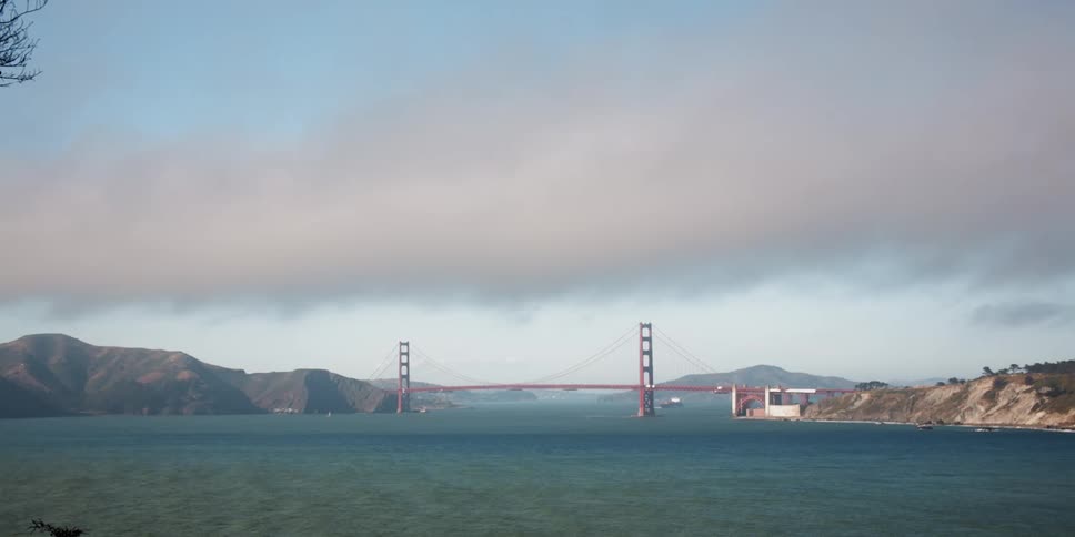 A suspension bridge over water and mountains in the distance under a cloudy sky