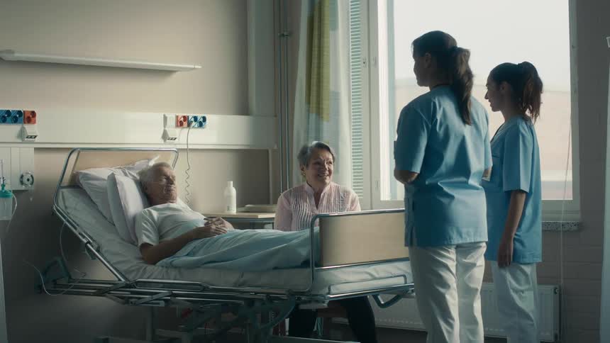 Medical personnel stand at foot of hospital bed talking to elderly patient and his visiting wife