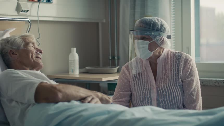 Visitor wearing face shield and mask holds hands with patient lying in hospital bed as they talk