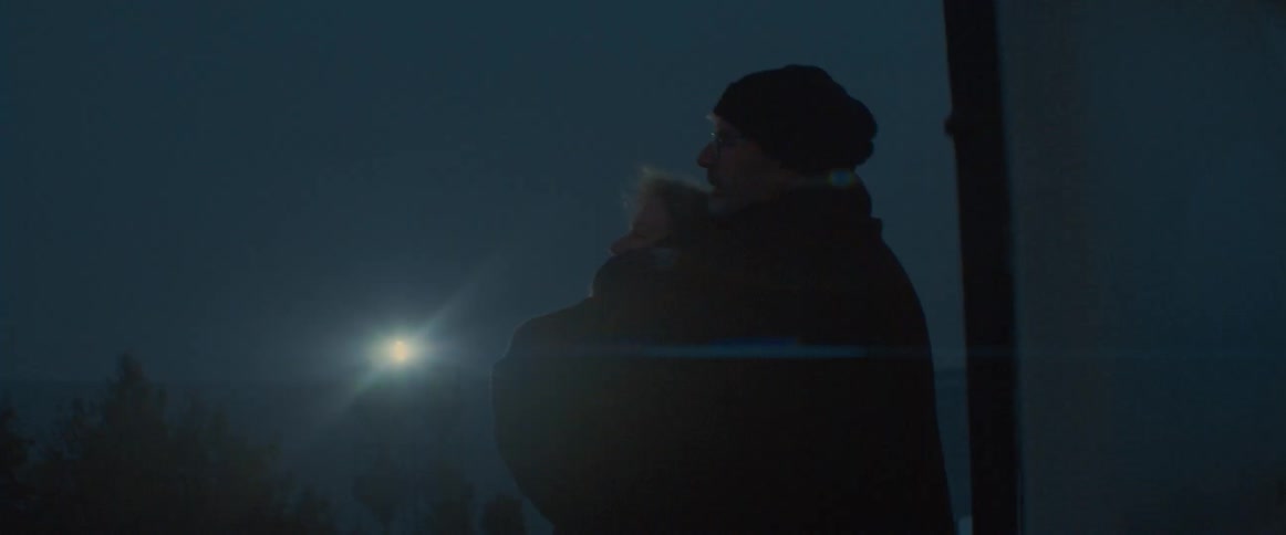 A man hugs his wife from behind as they stand near the coast