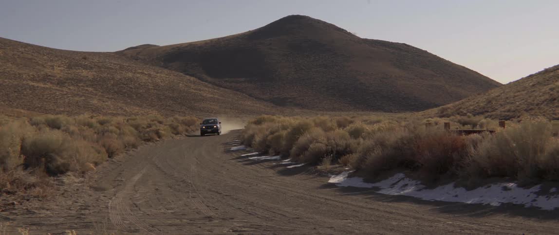 Black SUV driving along a dusty dirt road through a barren desert mountain landscape.