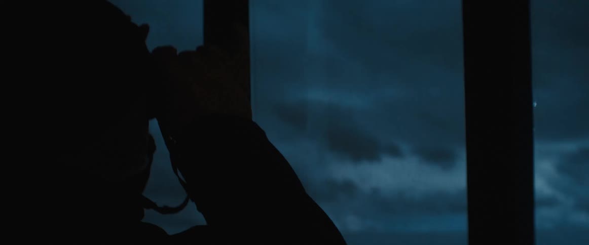 Man wears knit hat and coat standing next to beacon in lighthouse while he scans area with binoculars