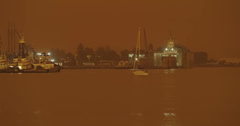 Sailboat motors across water passing ship sitting at dock on foggy amber day