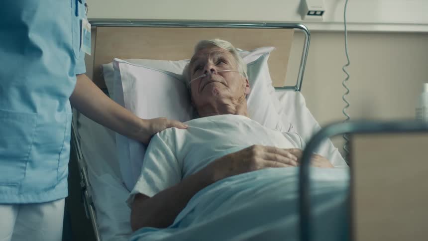 Old man lying in the hospital bed while a nurse approaches and touches him