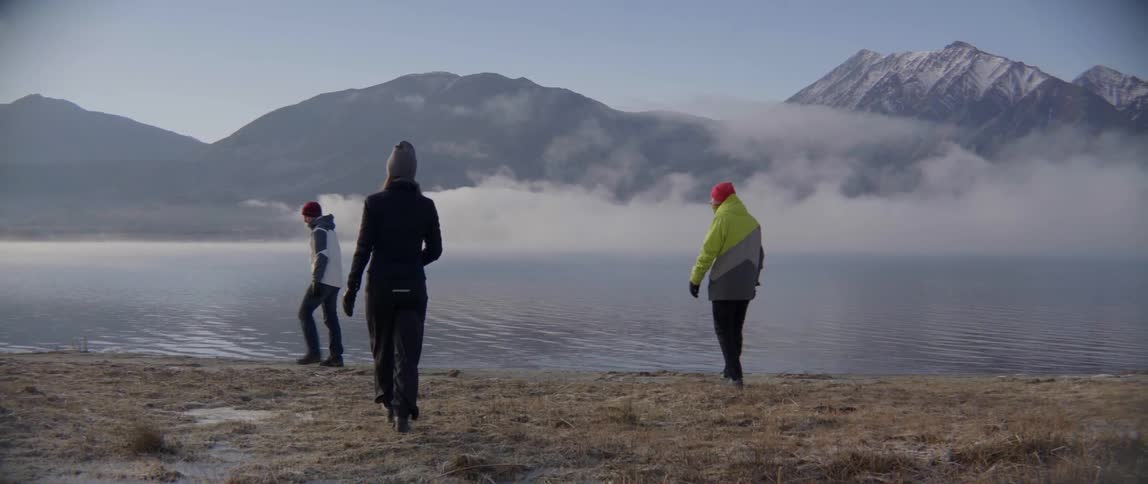 Three People Walking Toward the Water by a Mountain Range in a Quiet Outdoor Journey.