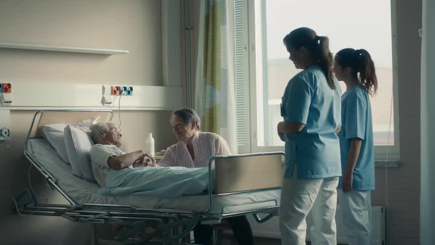 A pair of nurses talk to a patient and his wife in a hospital room before one nurse walks over to pat the shoulder of the patient
