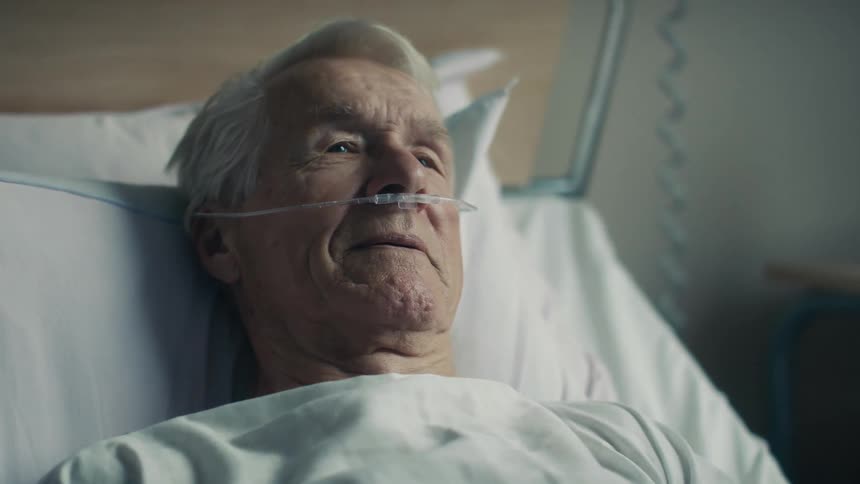 An elderly man lying in a hospital bed nods while talking to someone out of frame