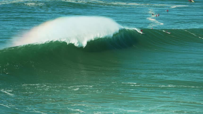 A large wave crashes on the ocean while surfers ride it