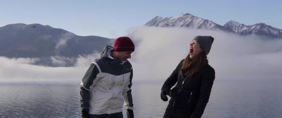 A man and woman laughing and standing in front of a lake with snow-covered mountains