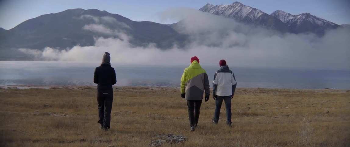 Three people walk across a field of brown grass toward a lake shrouded with clouds
