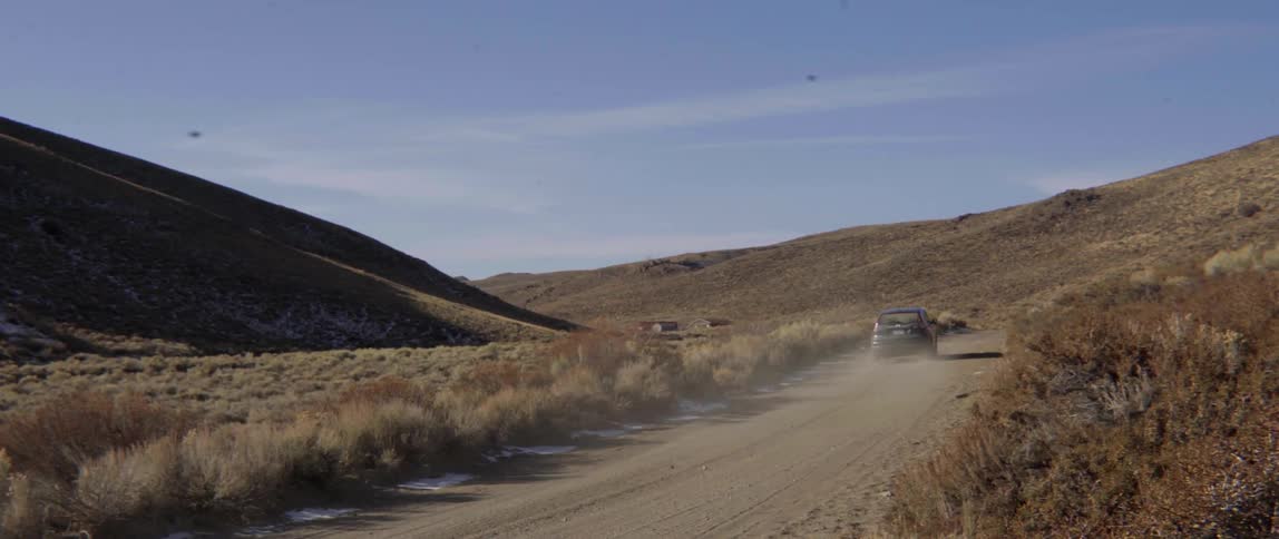 Car driving fast down a dusty dirt road in a vast barren mountain landscape.