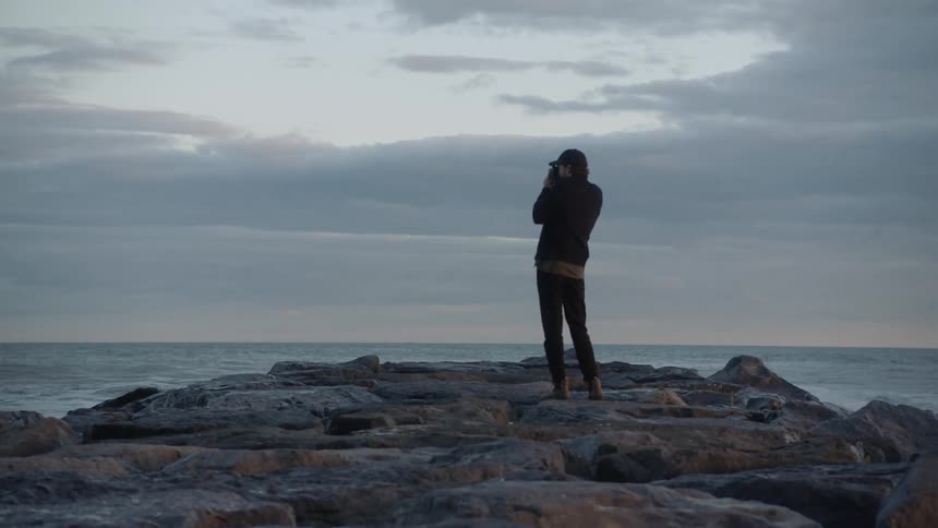 A man standing on a rocky shore lifts his camera to take a photograph as waves splash up