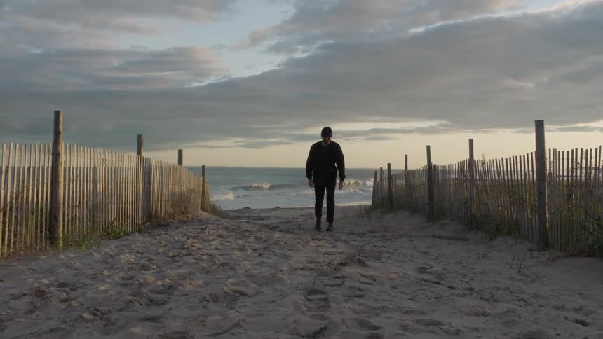 A man wearing black clothes is walking between two fences on a beach as the waves roll in behind him