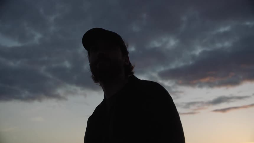 White man wearing a black baseball cap standing alone under the sunset looking around
