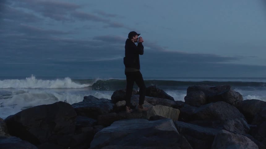 A photographer photographs the ocean while standing atop a pile of boulders on the coast in the evening
