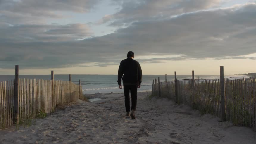 Man wearing baseball cap walks across sand between fences heading toward beach