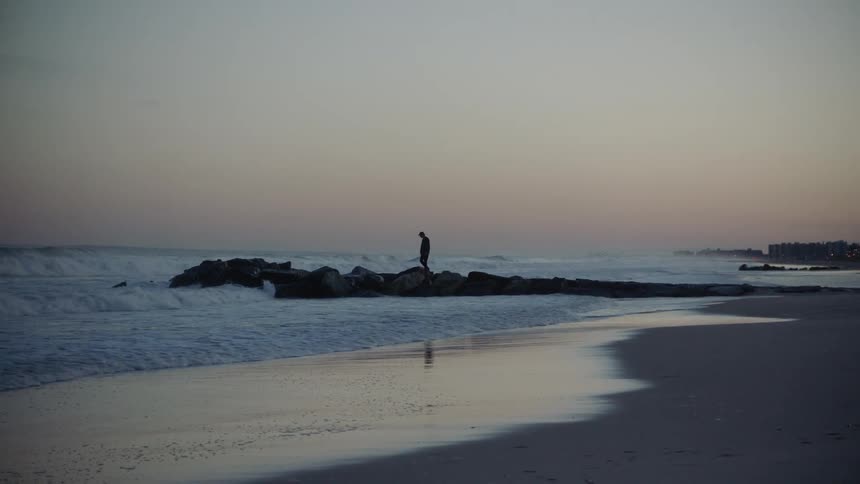 A man walks onto large boulders on the beach as strong waves wash onto the shore