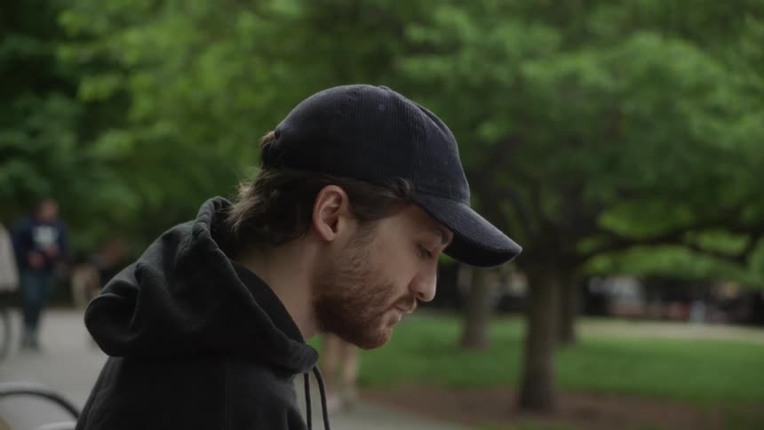 Man wearing a hoodie and baseball cap writing on a notebook sitting on a bench at a park