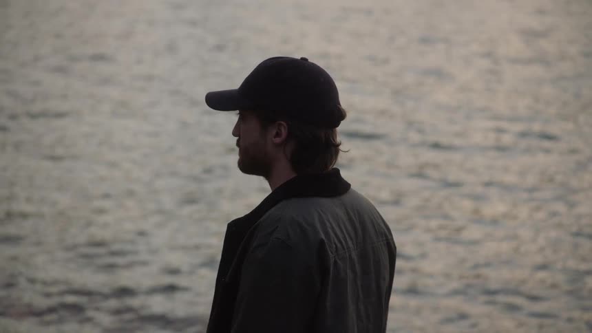 Bearded young man wearing a baseball cap looking at the surface of a lake at sunset