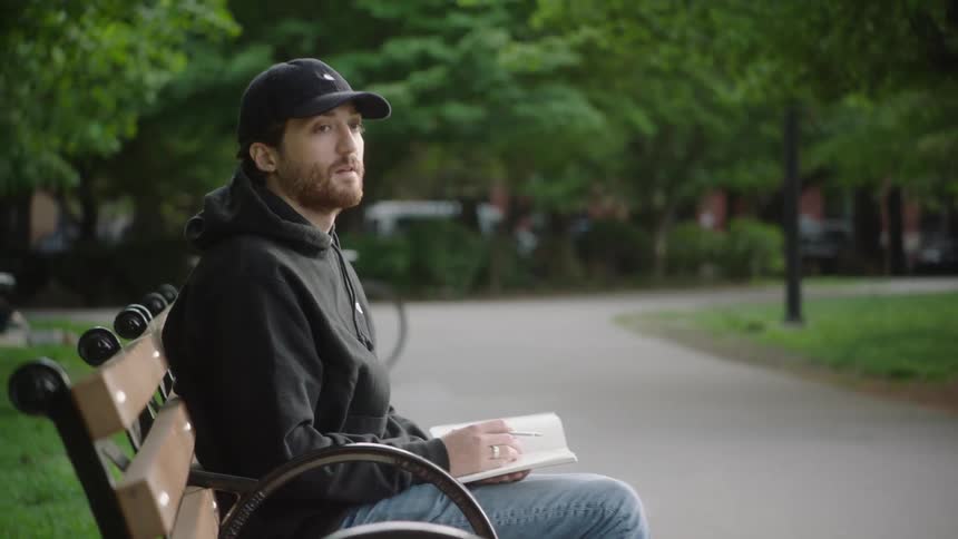 A man is sitting on a park bench holding a book and a pen as he looks around
