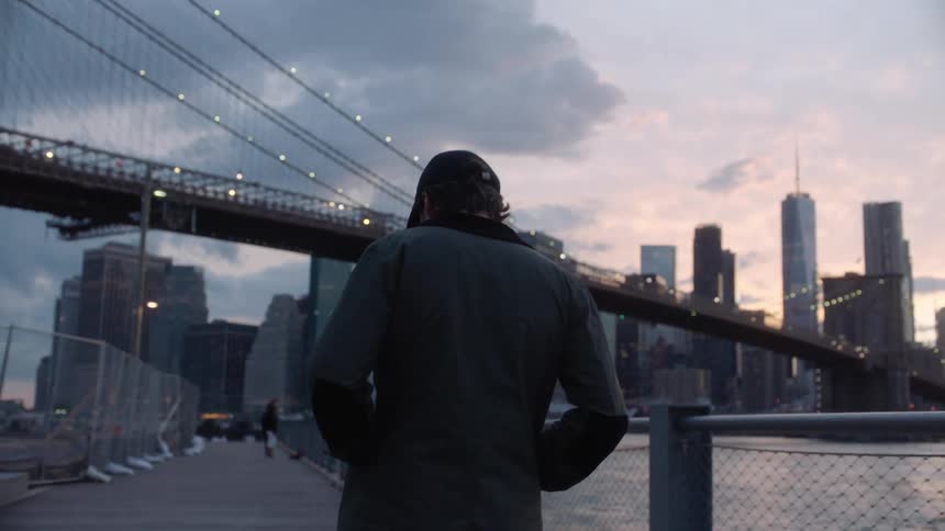 Pedestrian walks along boardwalk near water heading toward suspension bridge and downtowntower block