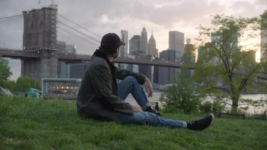 White man sitting at the grass next to the river looking to the city ahead
