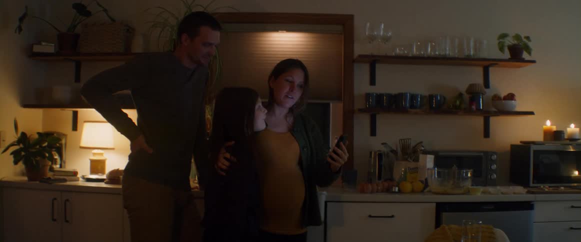 Expectant mother hugs daughter as she and family stand in kitchen talking via video call