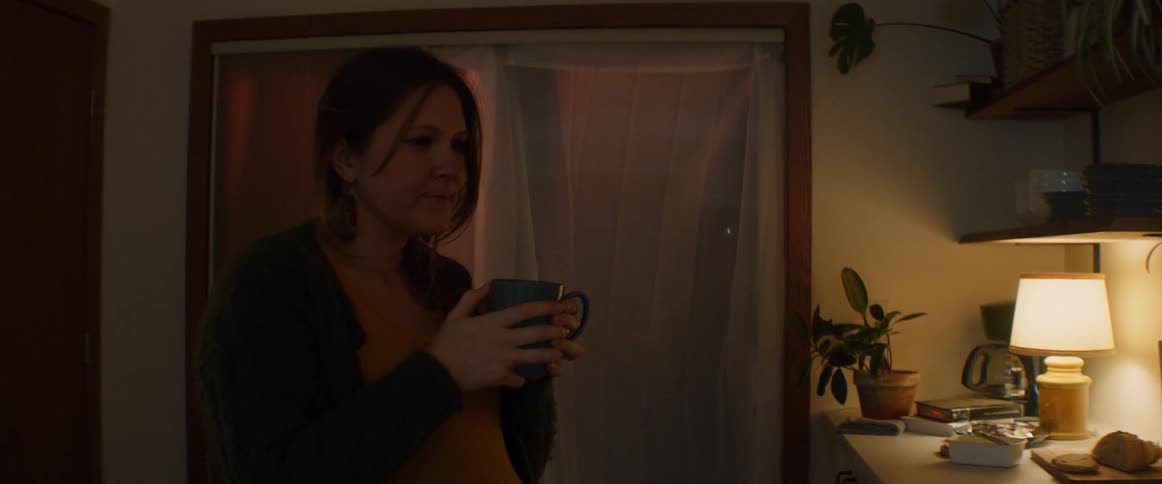Expectant mother stands in kitchen drinking from coffee cup and laughing as she listens