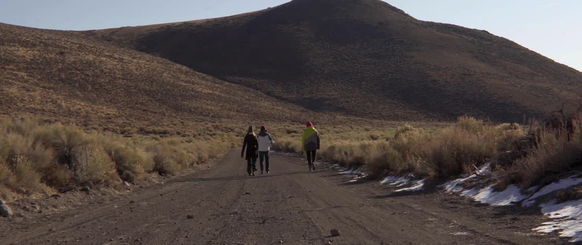 Three people hiking on dirt road toward rolling hills with energetic adventure.