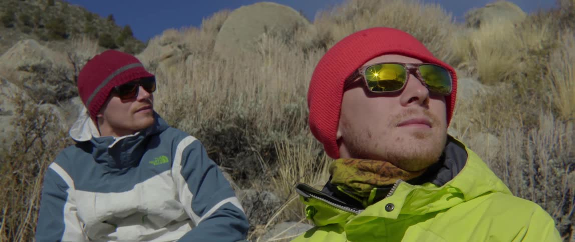 Two men wearing sunglasses sitting on hilltop with relaxed cool outdoor vibe.