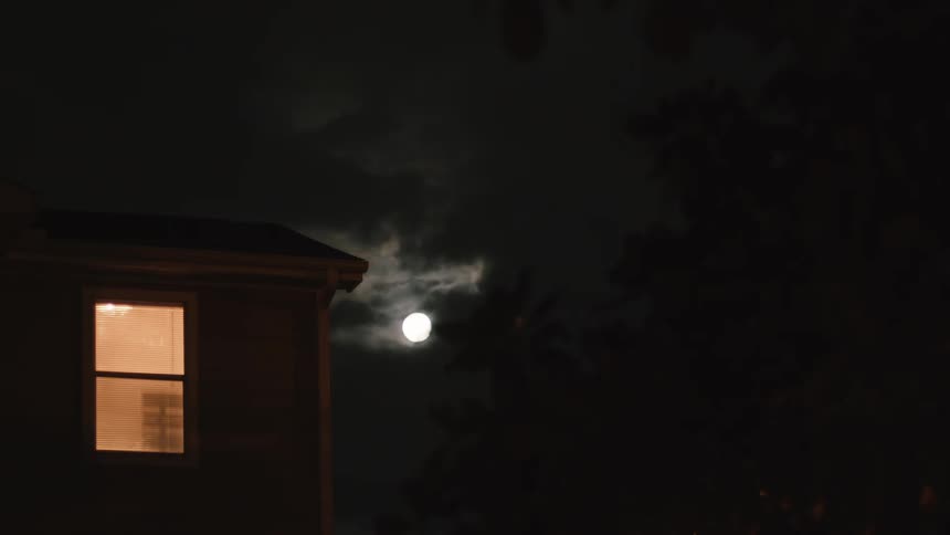 Clouds pass over a full moon in the night sky while a light shines in a house window