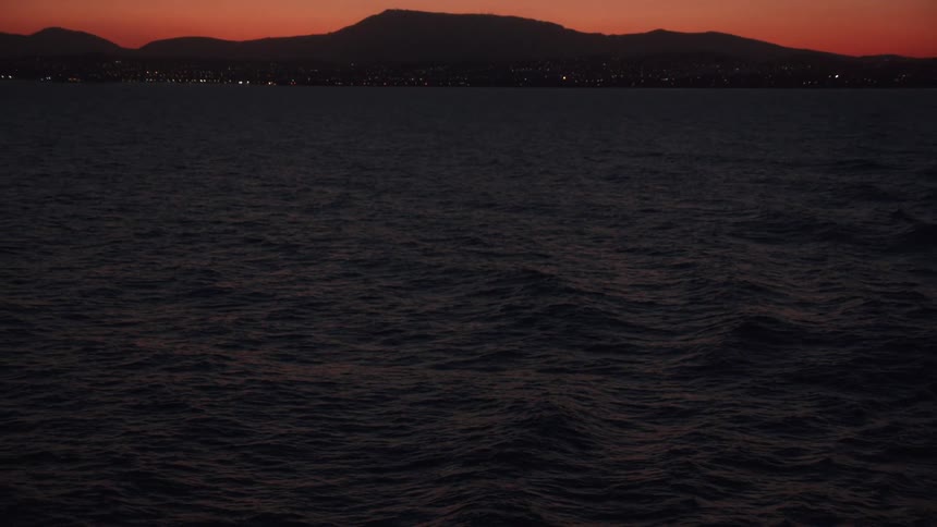 Waves roll across ocean as lights shine at base of mountains silhouetted against red sky