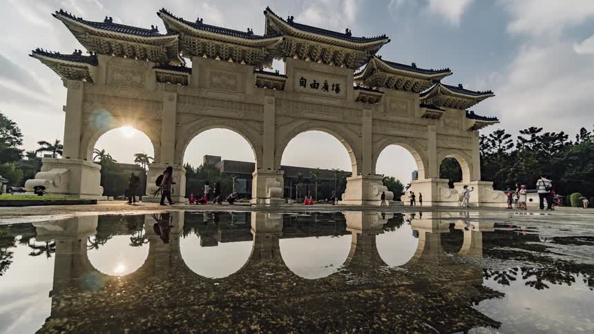 A large puddle reflects the liberty square arch in taiwan as tourists walk through