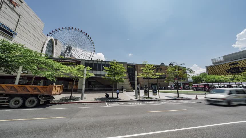Cars driving on a road next to a big mall in the city with a ferris wheel on the back