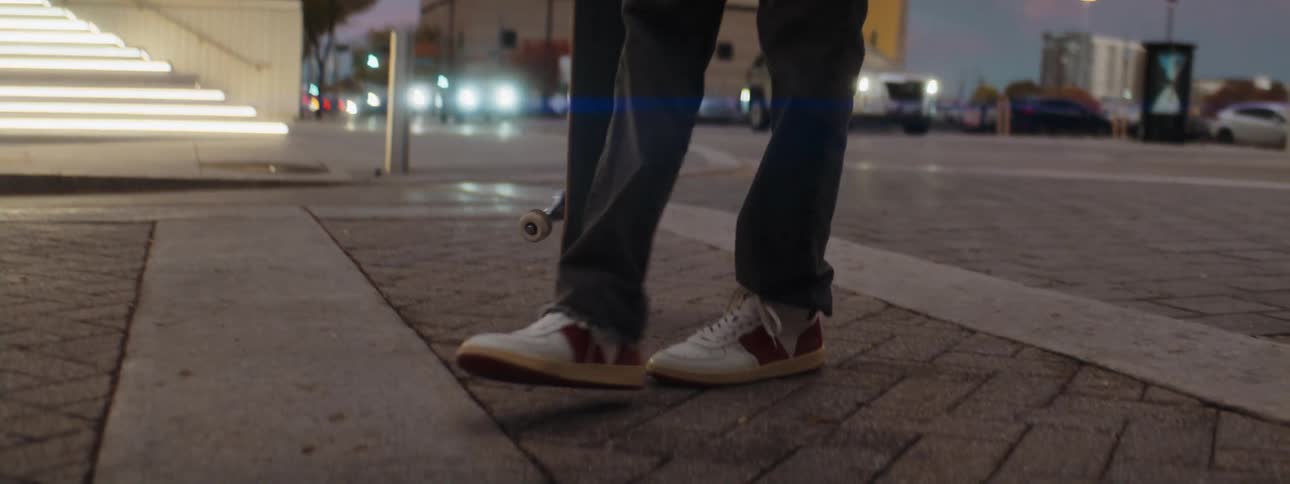 A male stands on a road and turns around as he picks his skateboard up by flipping it up with his foot as cars drive past