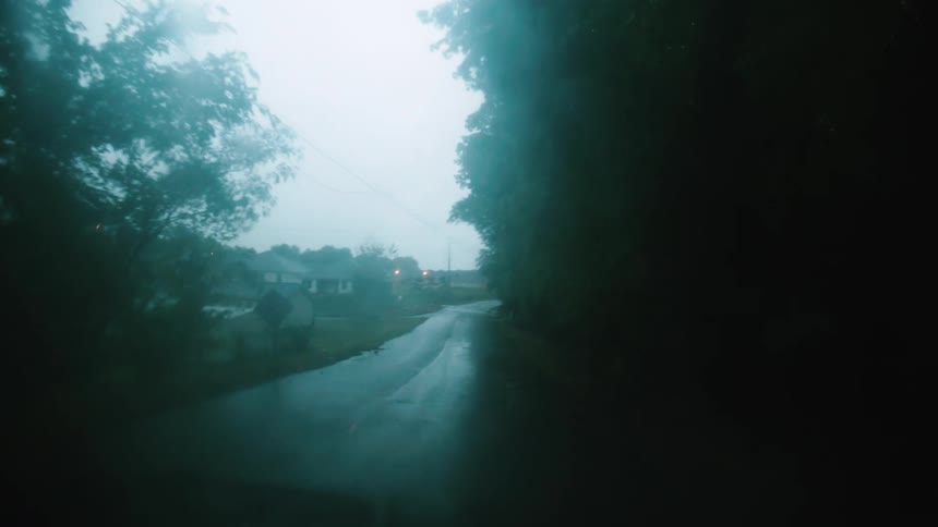 A car drives down a rainy street in a neighborhood lined with trees
