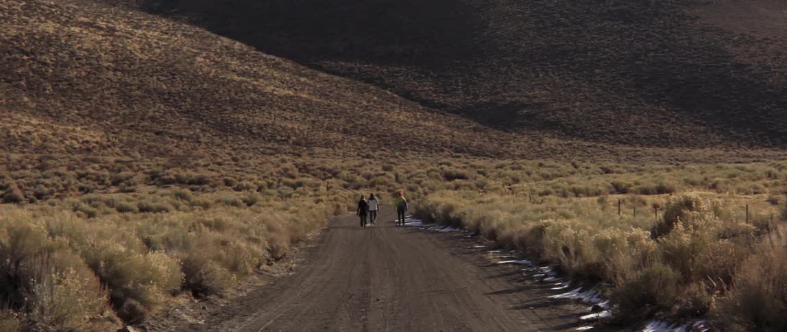 Three people are walking on a dirt road towards some hills