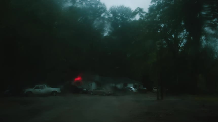 A fire burns at the back of a house in a rural area with cars parked in front