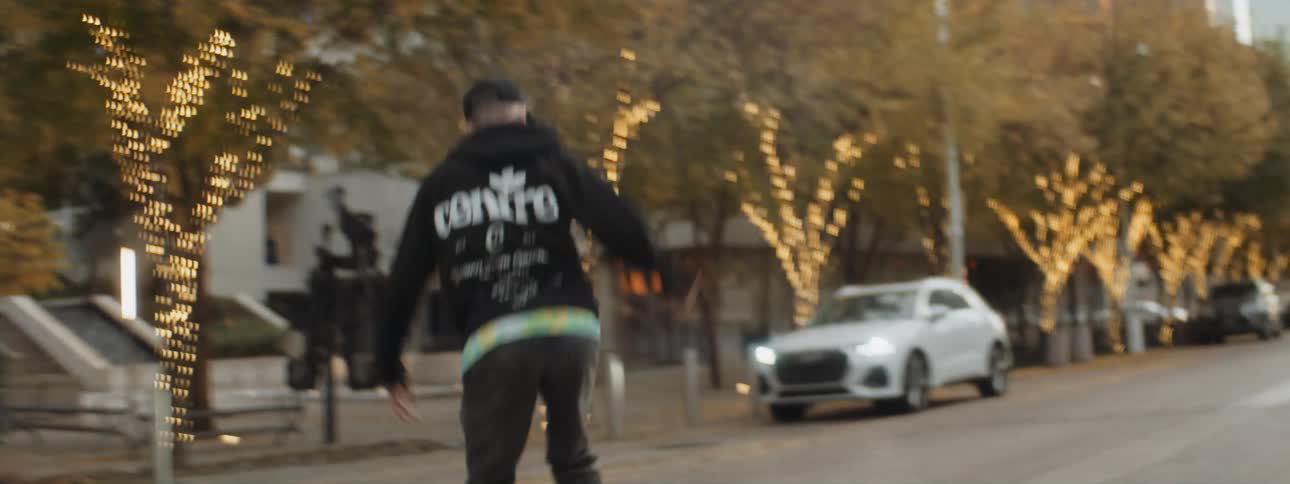 A man rides a skateboard down the center of a street past buildings and parked cars