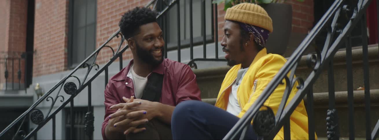 Two men sit on steps outside an apartment building and talk