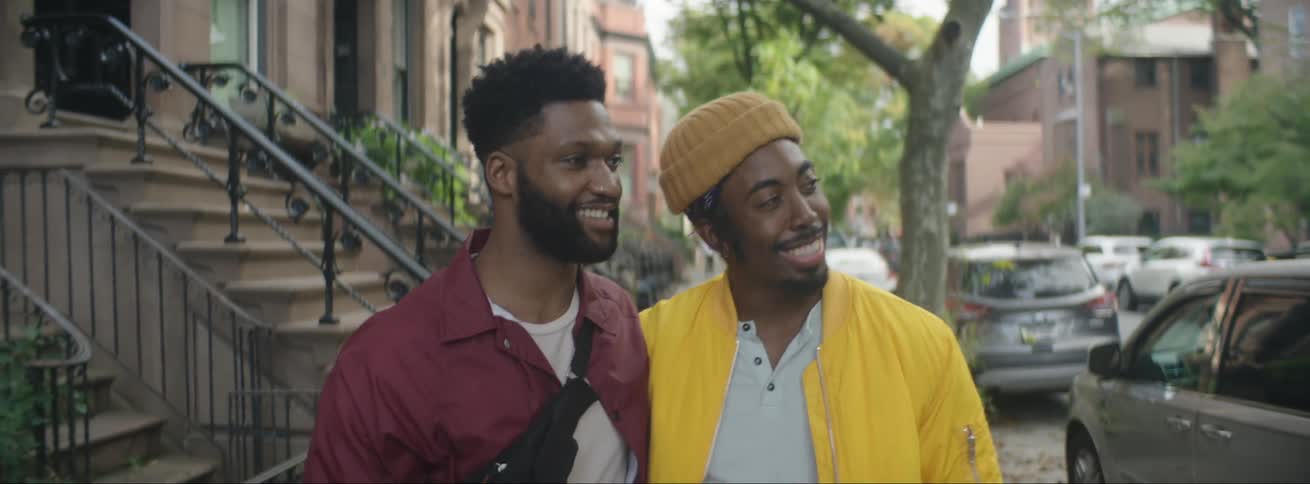 Two men walk down a neighborhood street with their arms around one another and talk as one man points and puts his hand on the other mans chest as cars drive past
