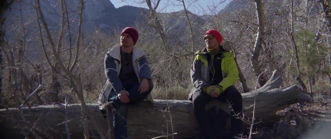 Two men sitting on log near mountain with calm reflective outdoor vibe.