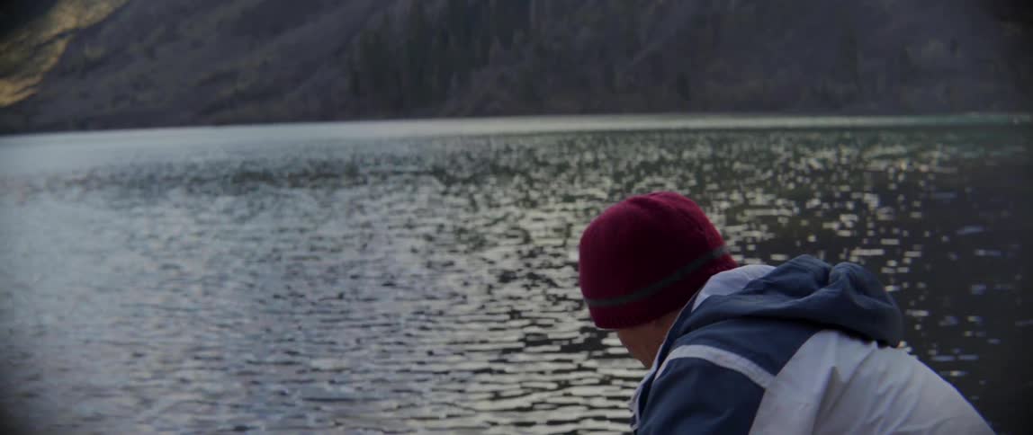 A man looks over to a mountain before crouching by the water before it