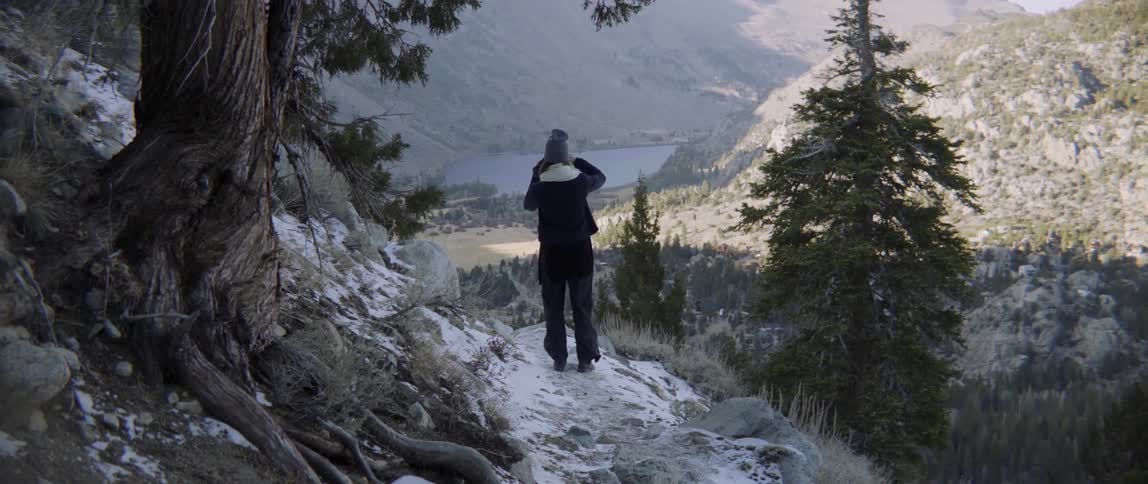 A person walks over some rocks photographing the hills and water