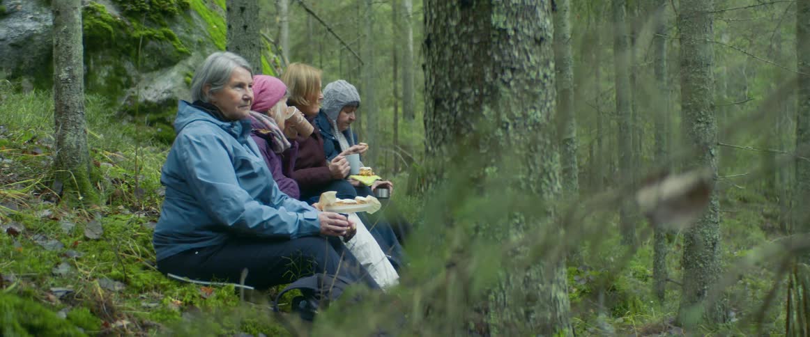 A woman and teenagers sit in a wooded area drinking from mugs