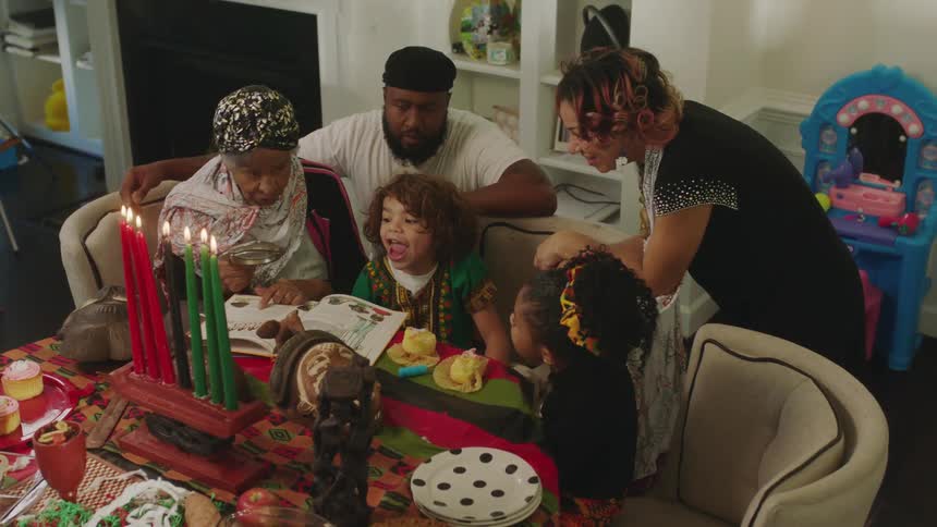 Grandma reads to her grandchildren as mom and dad stand close with kwanzaa candles lit on the table