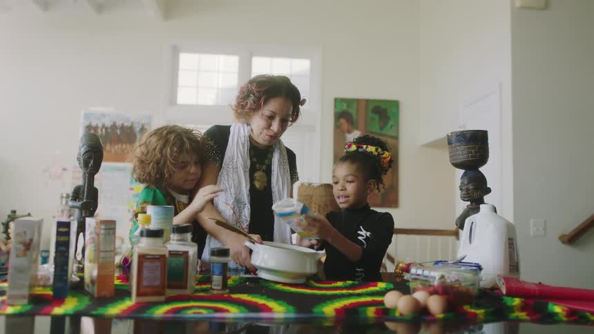 A woman helps young children pour food ingredients into a bowl