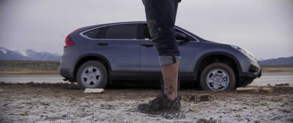 Person Standing by a Parked Car Looking Down at Muddy Shoes After a Walk.