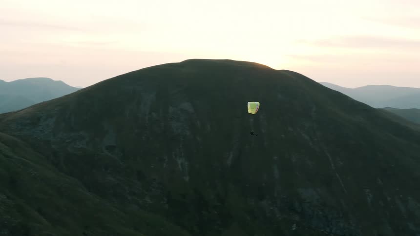 A person flies on a paraglider over rolling hills during sunset