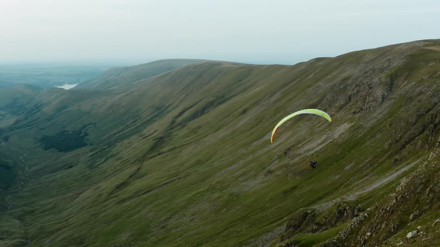 Person parasails through green valley toward foggy horizon